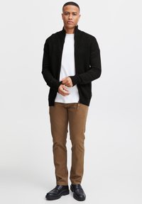 Schwarze Strickjacke mit hohem Kragen, über einem weißen T-Shirt, kombiniert mit braunen Chinos und schwarzen Schuhen. Verfügt über seitliche Reißverschlüsse.