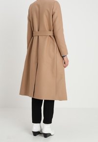 Dlouhý velbloudí kabát s páskem v pase, strukturovaný design a zadní rozparek. Nosený přes černé kalhoty a bílé kotníkové boty. Hladká textura.