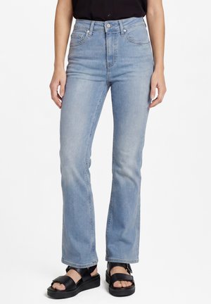 Femme portant un jean droit taille haute bleu clair et des sandales plateformes noires, debout devant un fond blanc.