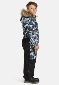 Dažādkrāsains sniega jaka ar kažokādas apdari uz kapuces, ar ziliem un melniem abstraktiem rakstiem; apvienota ar melnajām sniega biksēm un izturīgām melnām zābakām.