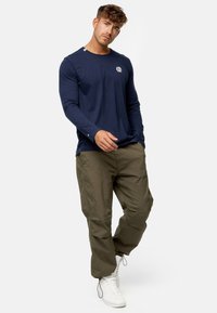 Camisa de manga larga azul marino con logo, combinada con pantalones cargo ligeros en color oliva y zapatillas altas blancas. Diseño simple y ajuste relajado.