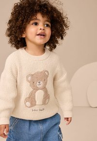 Maglione lavorato a maglia color crema con una grafica di un orso sul davanti, dotato di polsini a costine. Il bambino indossa pantaloni cargo in denim blu.
