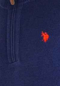 Marinefarbener Strickpullover mit einem Viertelreißverschluss, ausgestattet mit einem kleinen roten gestickten Logo auf der linken Brust sowie gerippten Bündchen und Kragen.