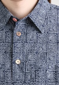 Marineblauwe shirt met witte abstracte patronen. Heeft een puntige kraag en twee lichtbruine knopen op de voorkant.