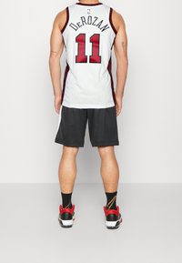 Homme debout de dos portant un maillot de basket blanc avec "DeRozan 11", un short noir, des chaussettes Nike noires et des baskets rouges et noires.