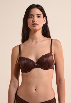 Mujer con cabello largo y oscuro, con un sujetador marrón con encaje y ropa interior a juego sobre un fondo beige.
