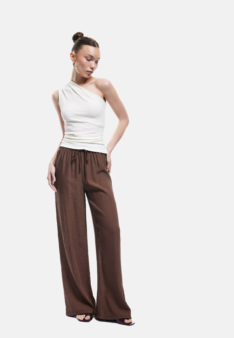 Top blanco de un solo hombro con tejido texturizado, combinado con pantalones amplios de crepé marrones que cuentan con una cinturilla elástica y detalles de patrón sutiles.