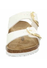 White two-strap sandal with gold buckles, cork footbed, and textured rubber outsole. Flat design suitable for casual wear.