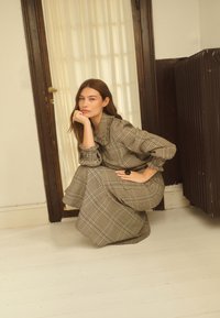 Femme en robe longue à carreaux accroupie, une main sur le menton et l’autre sur la hanche près d’une fenêtre et d’un radiateur sombre.