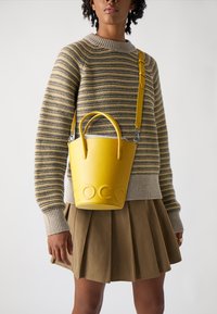 Sac seau en cuir jaune avec logo embossé, poignées arrondies et une bandoulière amovible, associé à un pull rayé et une jupe plissée.