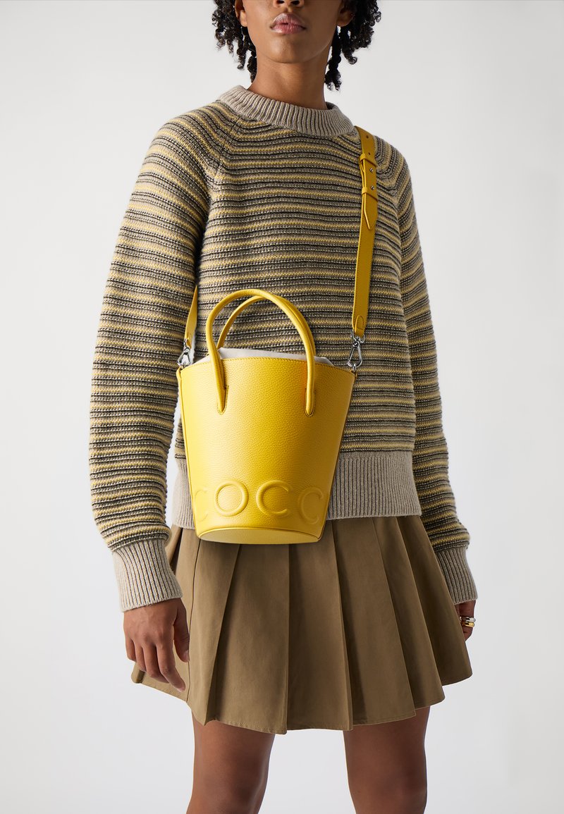 Sac seau en cuir jaune avec logo embossé, poignées arrondies et une bandoulière amovible, associé à un pull rayé et une jupe plissée.