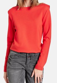 Maglietta lunga rossa in tessuto elasticizzato con dettagli sulle spalle, abbinata a jeans grigi con bottoni e tasche decorative.