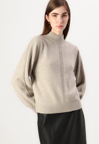 Beige stickad tröja med hög krage, ribbad fåll och detaljpaneler på sidan. Mjuk struktur och avslappnad silhuett, bärs över en mörk kjol.