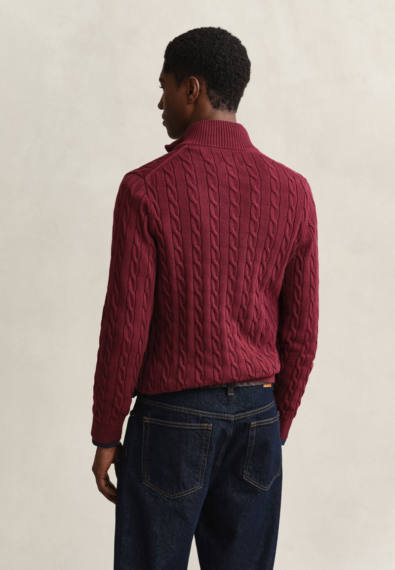 Man in a maroon cable knit sweater and dark blue jeans standing facing away against a plain beige background.