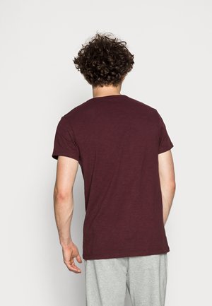 Mand med krøllet hår iført en ensfarvet vinrød t-shirt og lysegrå bukser, stående med ryggen mod en hvid baggrund.