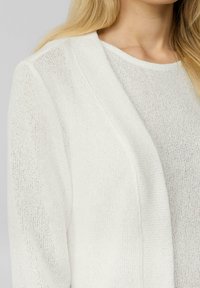 Weißer Strickcardigan mit offenem Frontbereich, ausgestattet mit einem strukturierten Stoff und einem runden Ausschnitt. Nahaufnahme der Ärmel und des Kragens.