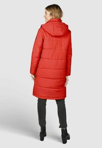 Veste matelassée rouge avec capuche, dotée de coutures horizontales, de fentes latérales et de détails en métal noir, assortie à un pantalon foncé et des bottes.
