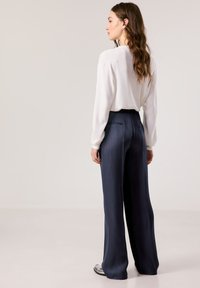 Weißes, langärmliges, Rundhalsoberteil kombiniert mit hochgeschnittenen, dunkelblauen, weiten Hosen. Die Hosen haben eine glatte Textur und Gesäßtaschen.