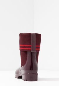 Bota de lluvia de goma color marrón con puño de punto doblado hacia abajo, que presenta dos franjas horizontales rojas y una asa en la parte posterior.