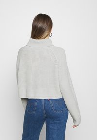 Virkat stickad tröja i ljusgrått med hög krage, långa raglansärmar och kortad fåll. Bärs tillsammans med blå denimjeans.