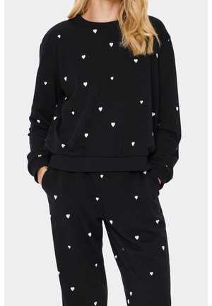 Kvinde iført sort sweatshirt med lange ærmer og bukser med små hvide hjerteprint, hænder i lommerne, blond hår synligt.