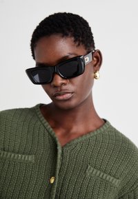 Čierne oversized slnečné okuliare s masívnym dizajnom a štvorcovým tvarom; majú jemný zlatý akcent po boku. Model má na sebe textúrovaný zelený kardigán.