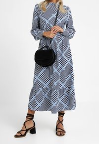 Långärmad klänning i svart och blå diamantsmönster, flödande tyg till vid vad, matched med svart rund axelväska och remsandaler.