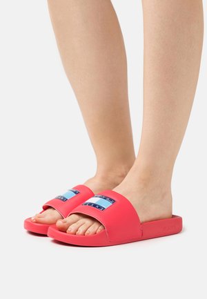 Chanclas de color rosa brillante hechas de material sintético suave. Presentan una correa ancha con un parche de logo azul y blanco. Diseño de suela plana.