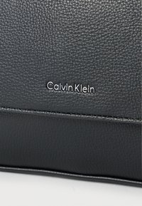 Μαύρο δερμάτινο προϊόν με υφή, με ασημένιο λογότυπο «Calvin Klein». Εντυπωσιακές ραφές κατά μήκος των άκρων.