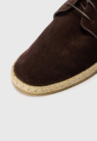 Pantofii din piele întoarsă maro au un vârf rotunjit, cu o talpă din jute. Textura este netedă, având șireturi și o margine a tălpii de o nuanță mai deschisă.