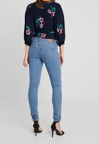Blusa oscura con estampado floral y mangas abullonadas, combinada con jeans ajustados de color azul claro y zapatos de tacón negros, exhibiendo una silueta ajustada.