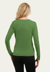 Femme à cheveux blonds longs portant un pull vert ajusté à manches longues et un pantalon noir, vue de dos sur un fond uni.