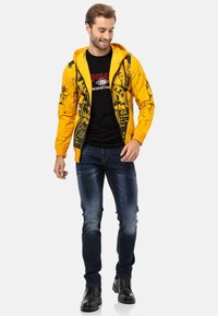 Haut à capuche zippé jaune avec imprimé graphique noir, porté sur un t-shirt noir. Un pantalon slim et des bottes noires à lacets complètent la tenue.