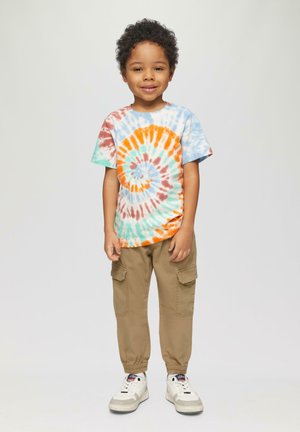 Katoenen tie-dye T-shirt met een oranje, blauwe en groene spiraalpatroon; beige cargobroek met zijzakken; witte sneakers met grijze accenten.