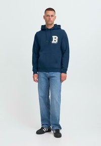 Jeune homme debout portant un sweat à capuche bleu marine avec un grand "B", un jean bleu clair et des baskets noires avec des rayures blanches sur un fond uni.