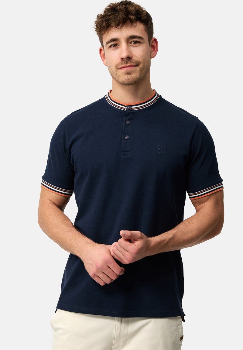 Marineblå polo T-shirt med korte ærmer, tre knapper ved kraven og stribede ribbede detaljer i rød, hvid og blå på halsudskæringen og ærmerne.