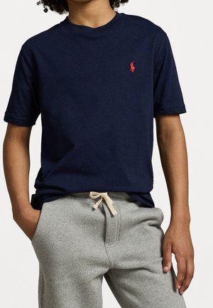 Jeune personne portant un t-shirt bleu marine à manches courtes avec un petit logo rouge et un pantalon de survêtement gris clair avec un cordon blanc.