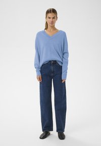 Világoskék, túlméretezett V-nyakú pulóver, sötétkék, magas derekú, egyenes szárú farmerrel és fekete, lapos, bebújós cipővel párosítva.