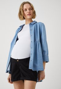 Lyseblå denimskjorte med trykknapper, avslappet passform og oppbrettede ermer, brukt over en hvit tanktopp og svarte skreddersydde shorts.
