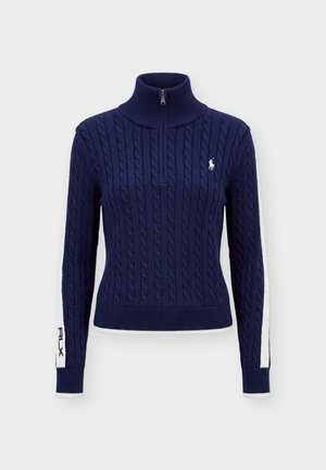Personne portant un pull torsadé bleu marine avec des rayures blanches et un logo, un pantalon large bleu marine et des baskets blanches, debout devant un fond uni.