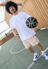Hvid Nike T-shirt med et farvet logo, lyserøde mesh shorts med hvide striber og grå-blå sneakers på en basketballbane. Basketball i hånden.