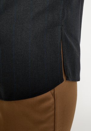 Zwart herringbone stoffen overhemd met blauwe strepen, voorzien van een zijsplit. Gecombineerd met gladde bruine stoffen broek, met strakke lijnen en snitten.