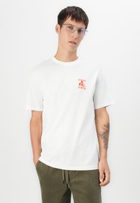 White cotton t-shirt featuring a small orange graphic with the text "SHANGHAI CHAMPION" on the left chest. Model wearing olive green pants.