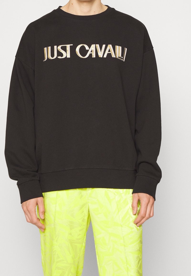 Personne portant un sweat-shirt noir avec le texte "JUST CAVALLI" en doré métallique et un pantalon à motifs jaune néon sur un fond uni.