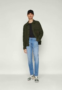 Jovem a usar um boné preto, uma jaqueta verde escura, uma camisa preta, jeans azuis claros e ténis brancos e verdes, a posar contra um fundo simples.