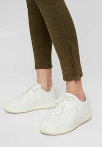 Jambes portant un pantalon slim vert olive avec des fermetures éclair aux chevilles, associées à des baskets à plateforme blanches à lacets sur un fond clair uni.
