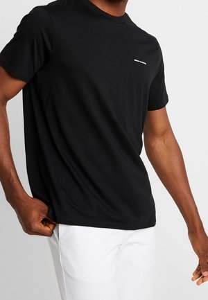 T-shirt noir en coton avec un col rond, des manches courtes et un petit logo blanc sur la poitrine, porté avec un pantalon blanc.