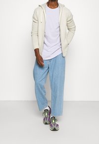 Felpa zip grigia chiara con coulisse, t-shirt lavanda, jeans in denim blu larghi e sneaker grigio-verdi con accenti viola.