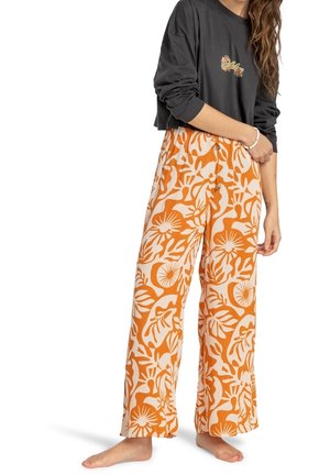 Femme portant un crop top noir à manches longues et un pantalon large taille haute avec un motif floral orange et beige audacieux, pieds nus, debout.