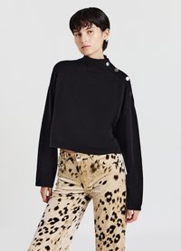 Schwarzer kurzer Pullover mit hohem Kragen und gerippten Bündchen, versehen mit drei dekorativen Metallknöpfen auf der linken Schulter. Kombiniert mit beigefarbenen Hosen mit Leopardenmuster.